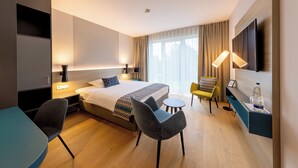 A modern hotel room with a bed, two chairs, a small table, a TV, and a bedside table with a lamp. - Parkhotel Maximilian Ottobeuren Resort&Spa (Ottobeuren)