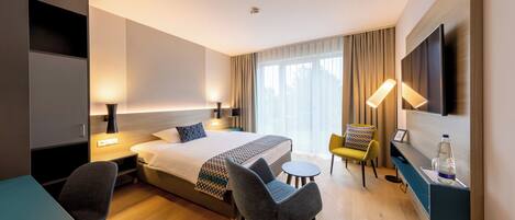Una habitación de hotel moderna con cama, dos sillas, una mesita, televisión y una mesita de noche con lámpara.