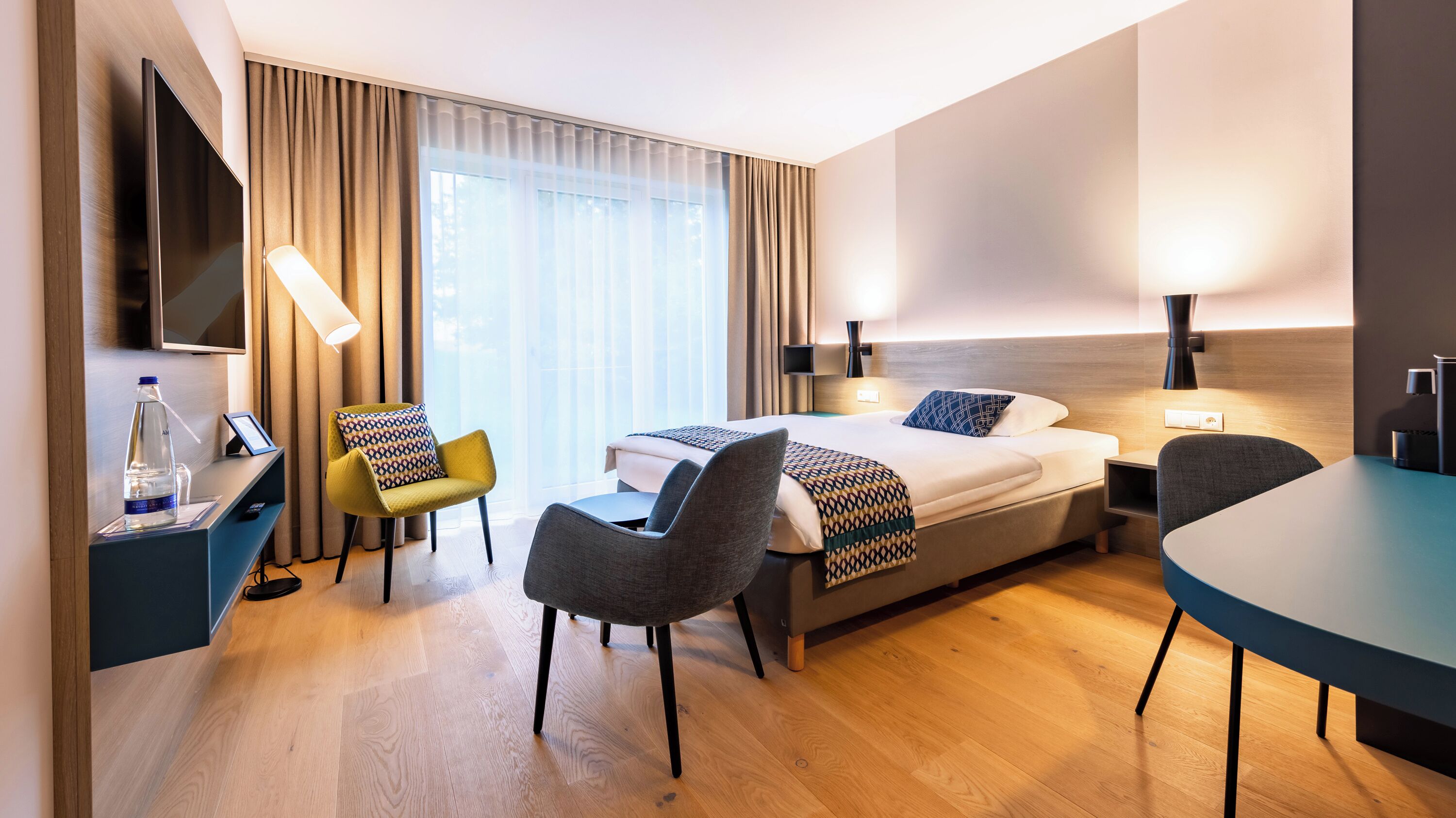 A modern hotel room with a bed, two chairs, a desk, a TV, and a large window with curtains.