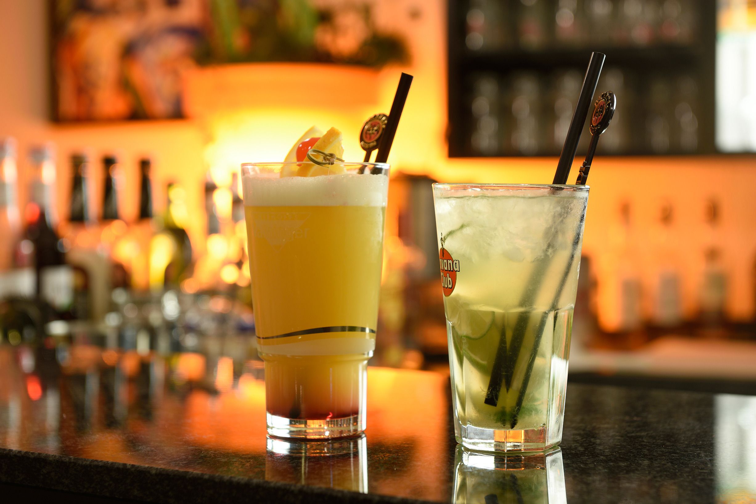 Two cocktails on a bar counter with a blurred background of bottles.