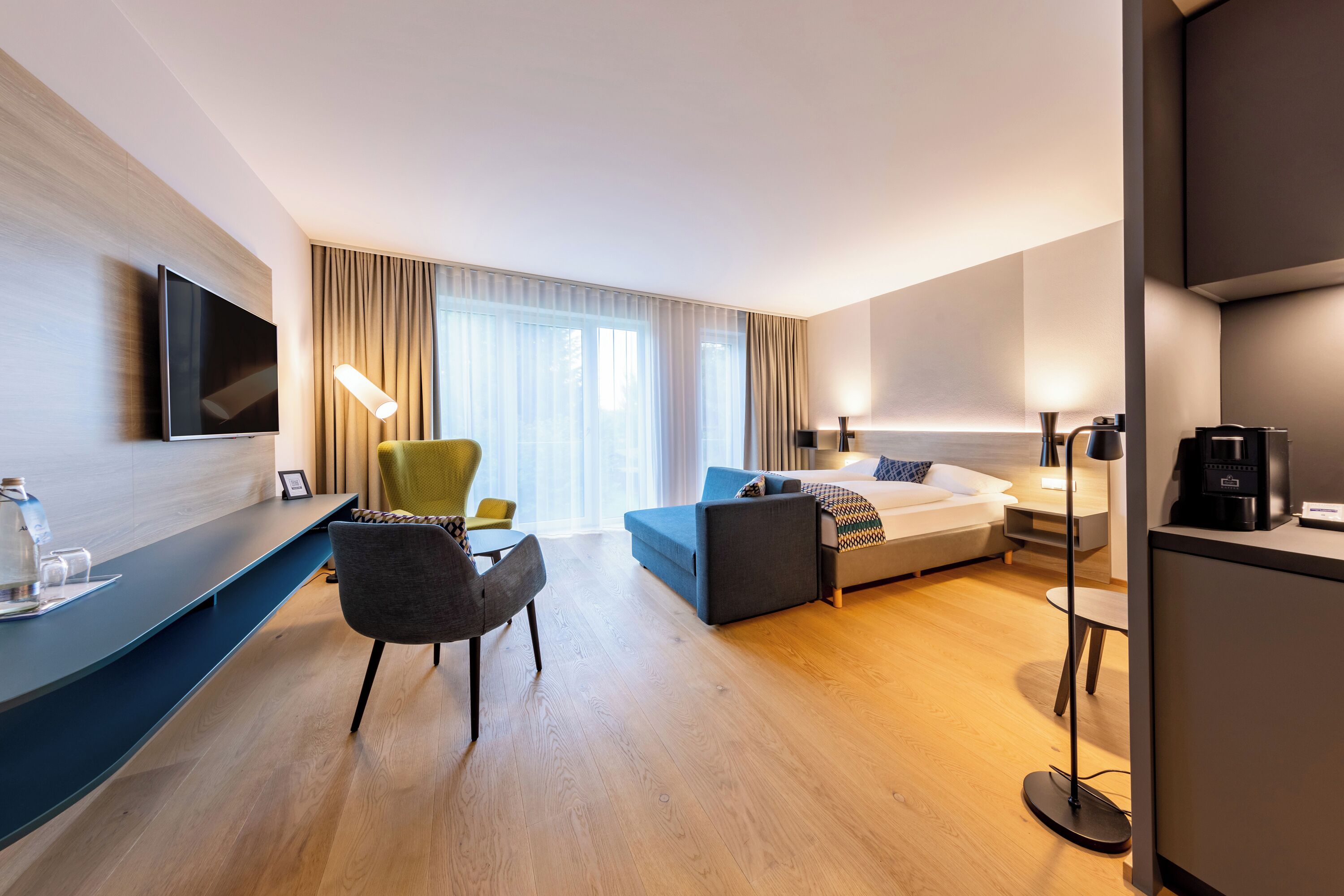 A modern hotel room with a flat-screen TV, a sofa, a desk with a lamp, a coffee machine, and a large window with curtains.