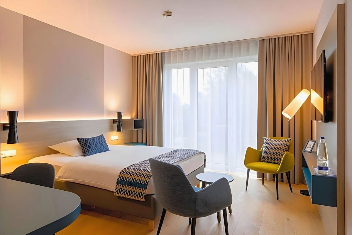 A hotel room with a bed, two chairs, a small table, a TV, and a window with curtains.