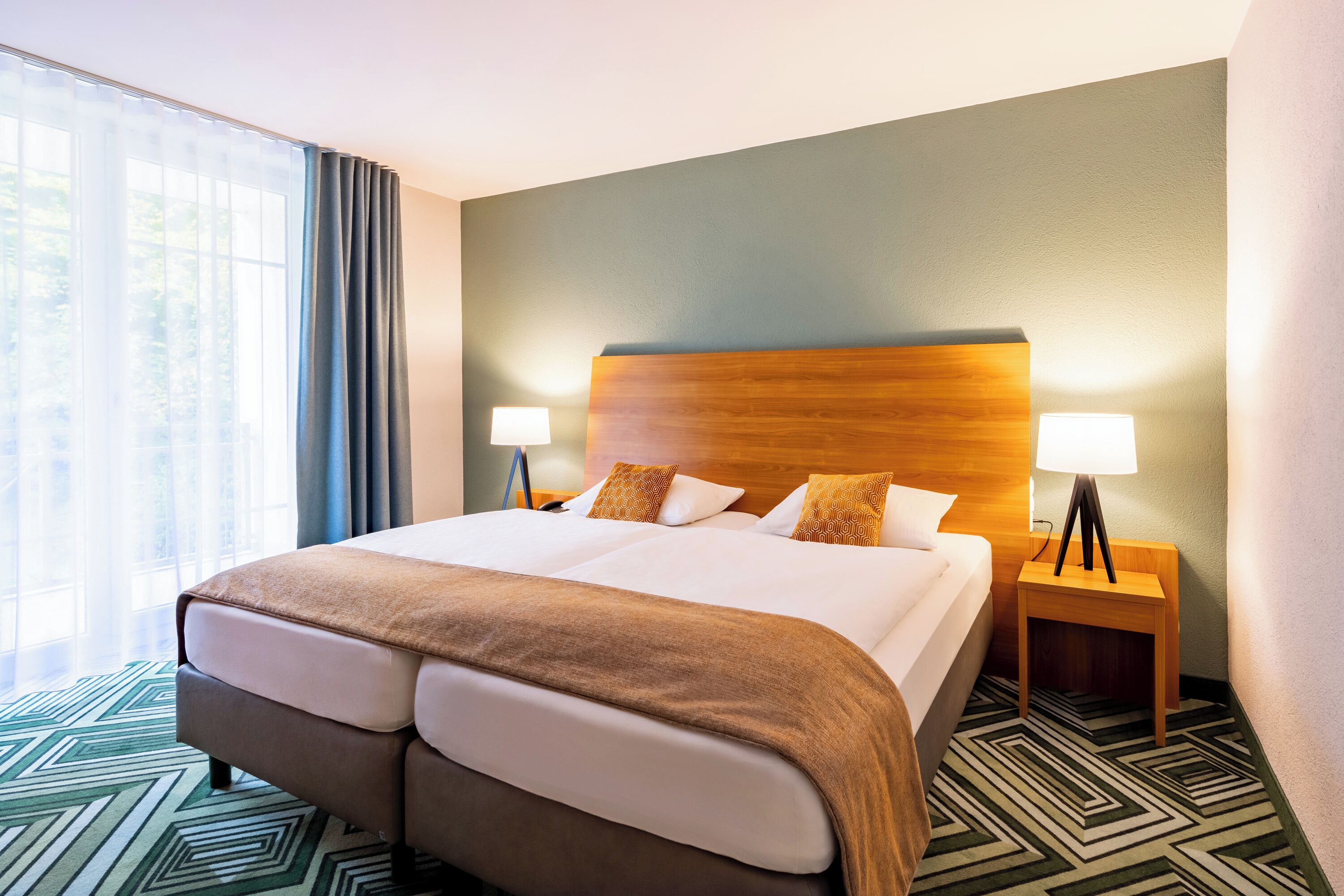 A hotel room with a large bed, two bedside tables with lamps, a wooden headboard, and a patterned carpet.
