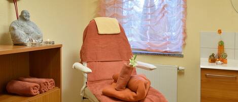 A massage chair with a towel and a decorative item on it, a statue on a shelf, and a window with sheer curtains.