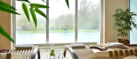A room with two reclining chairs, a small table with a vase and glasses, and a large window overlooking a garden.