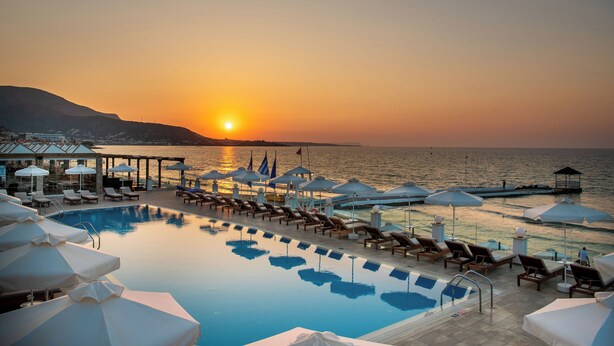 Un espace aménagé au bord de la piscine, avec des chaises longues et des parasols, offrant une vue sur une étendue d’eau au coucher du soleil.
