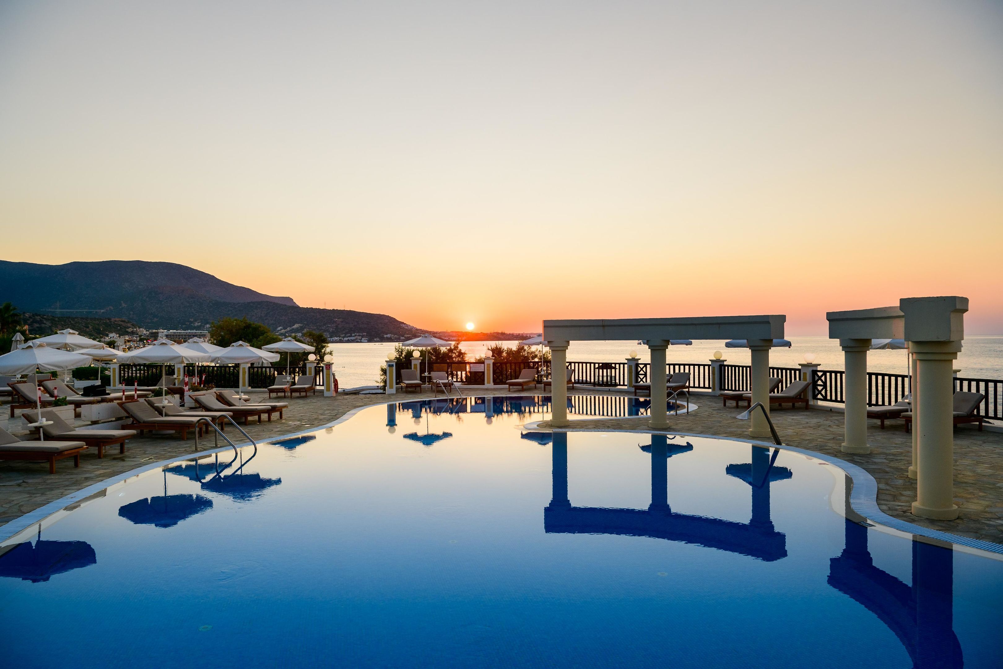 Ein Poolbereich mit Liegestühlen und Sonnenschirmen, mit Blick auf ein Gewässer und Berge bei Sonnenuntergang.