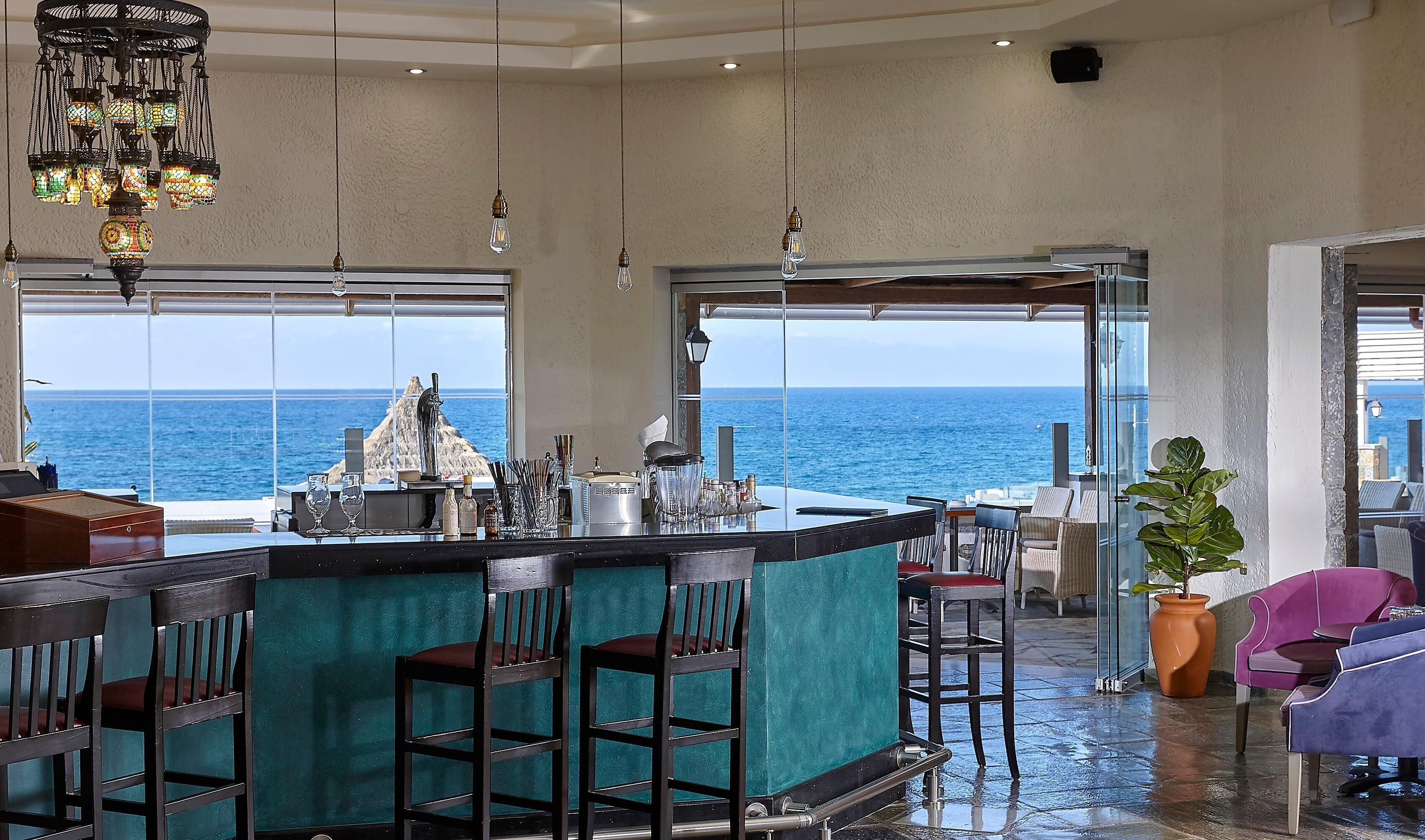 A bar with a view of the ocean, bar stools, and a large potted plant.