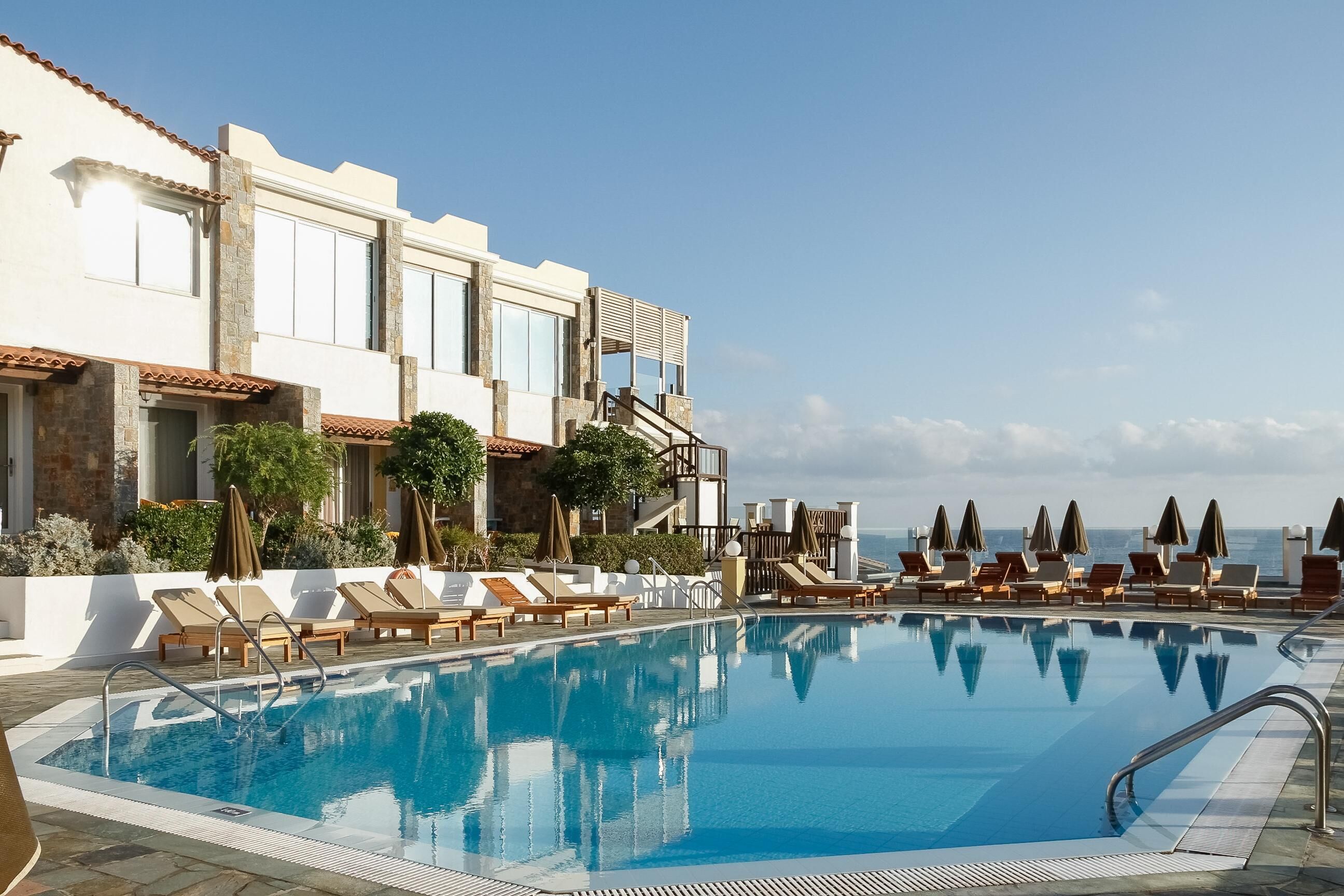 A swimming pool with lounge chairs and umbrellas, a building with stone and stucco exterior, and a clear sky.
