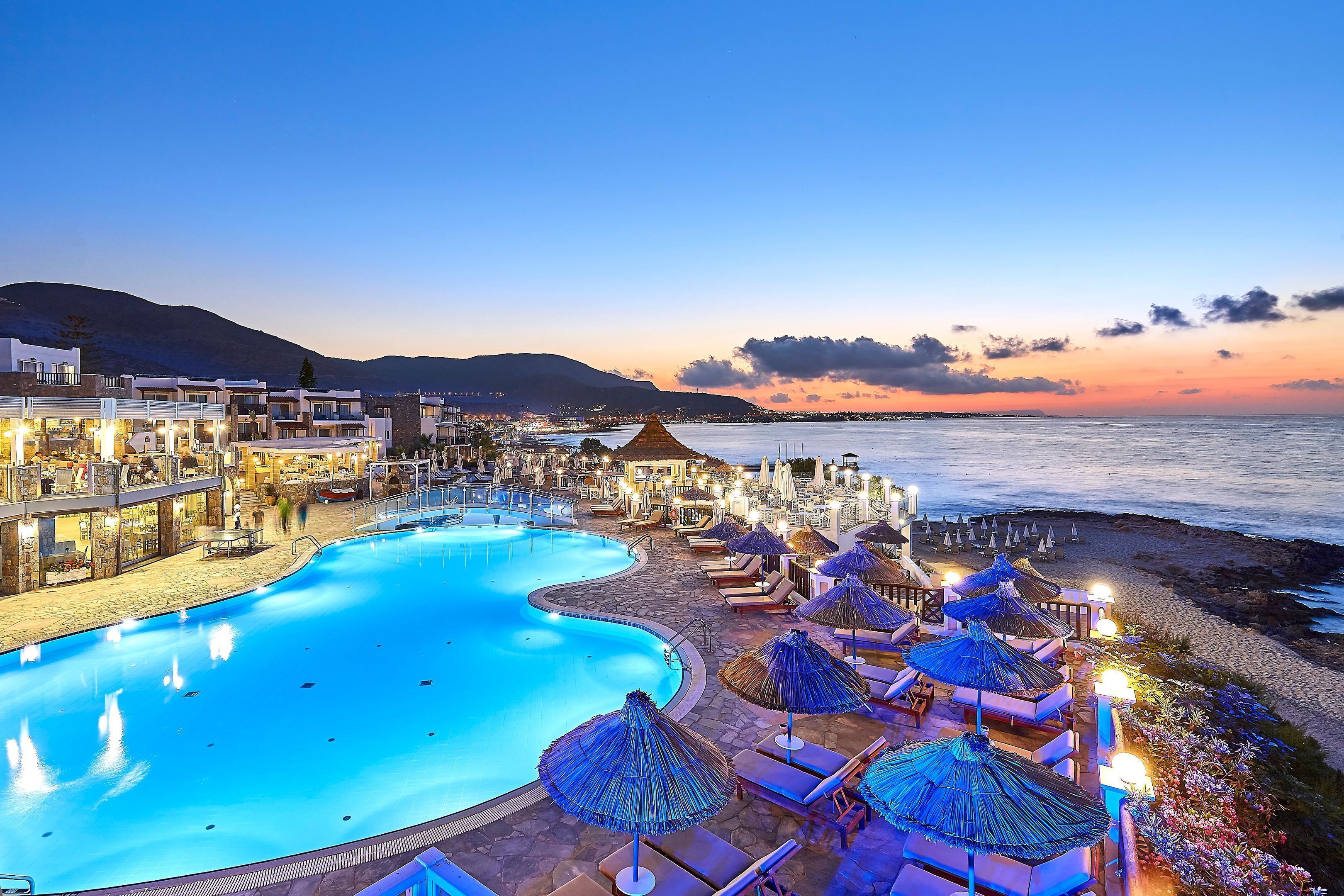 Un resort con piscina, zonas de descanso frente a la playa y vistas al océano al atardecer.