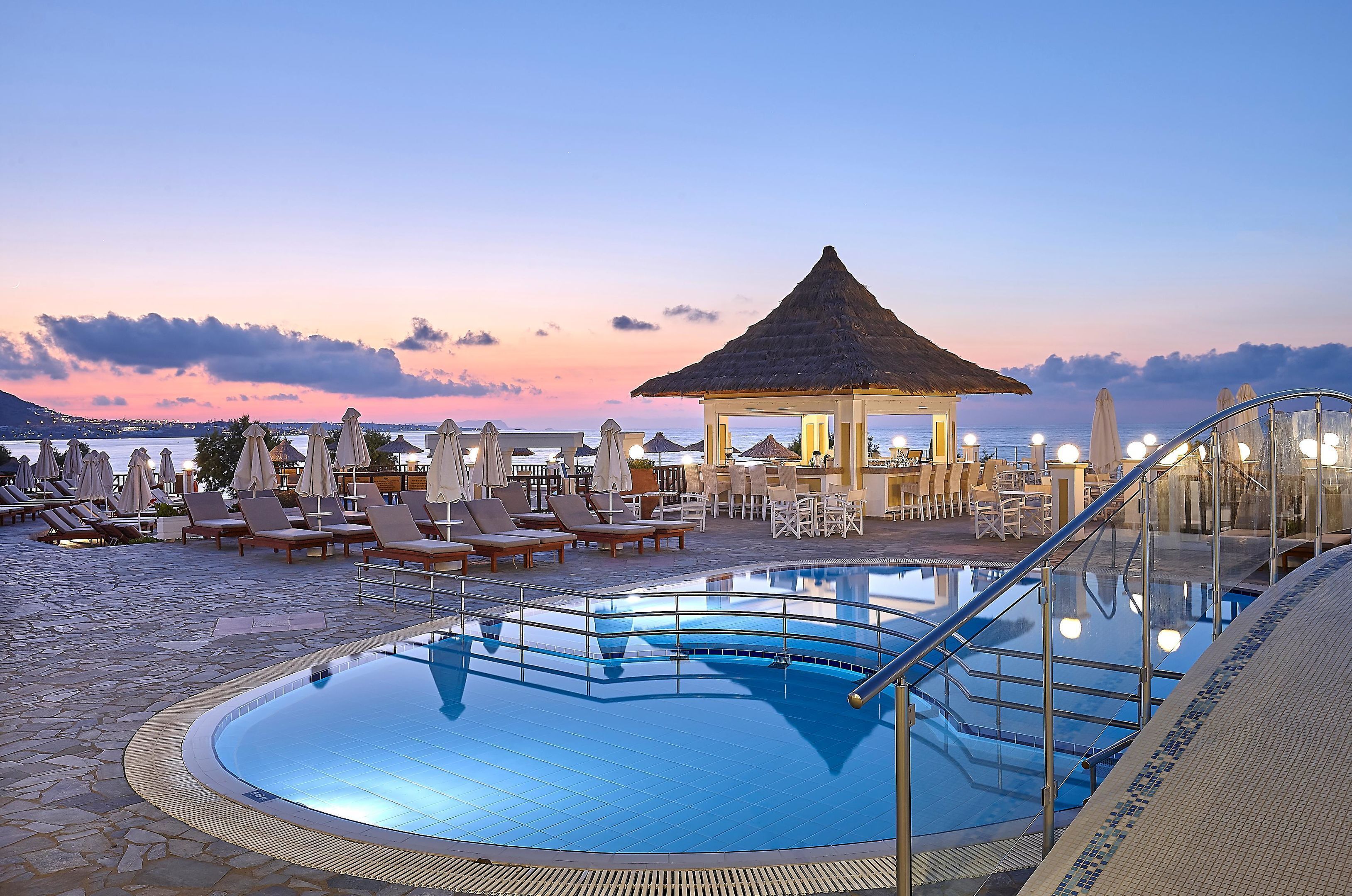A pool area with lounge chairs and umbrellas, a thatched-roof gazebo, and a clear view of the sea at sunset.