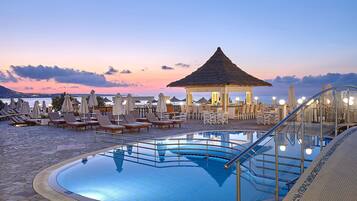 A pool area with lounge chairs and umbrellas, a thatched-roof gazebo, and a clear view of the sea at sunset.