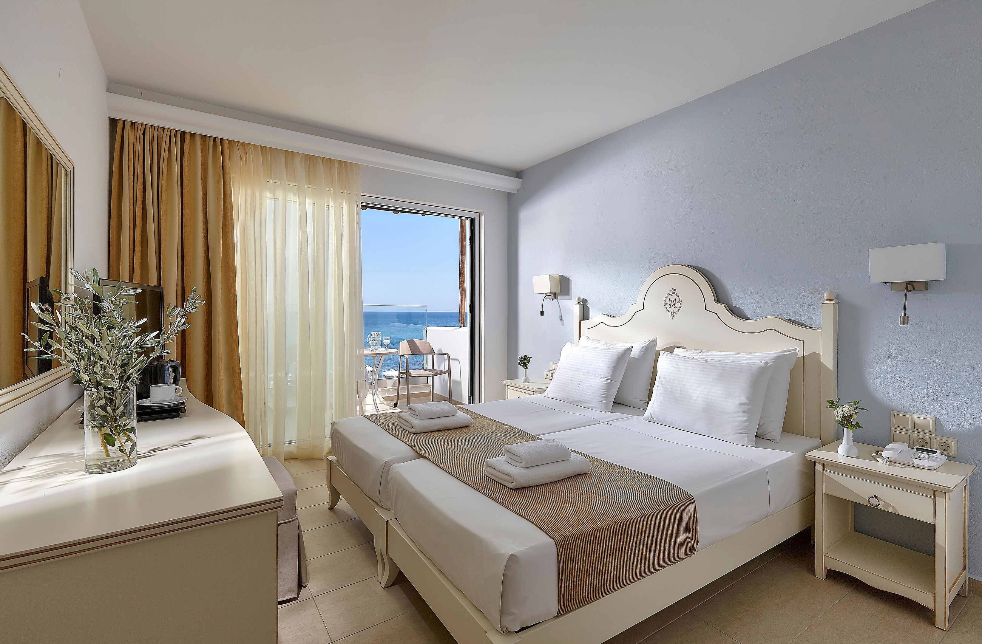 A hotel room with a bed, bedside tables, a desk with a vase, and a view of the sea.