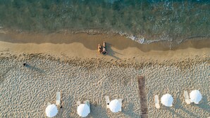 An aerial view of a beach with two people sunbathing, several beach umbrellas, and a wooden structure. - Alexander Beach Hotel & Village Resort (Hersonissos)