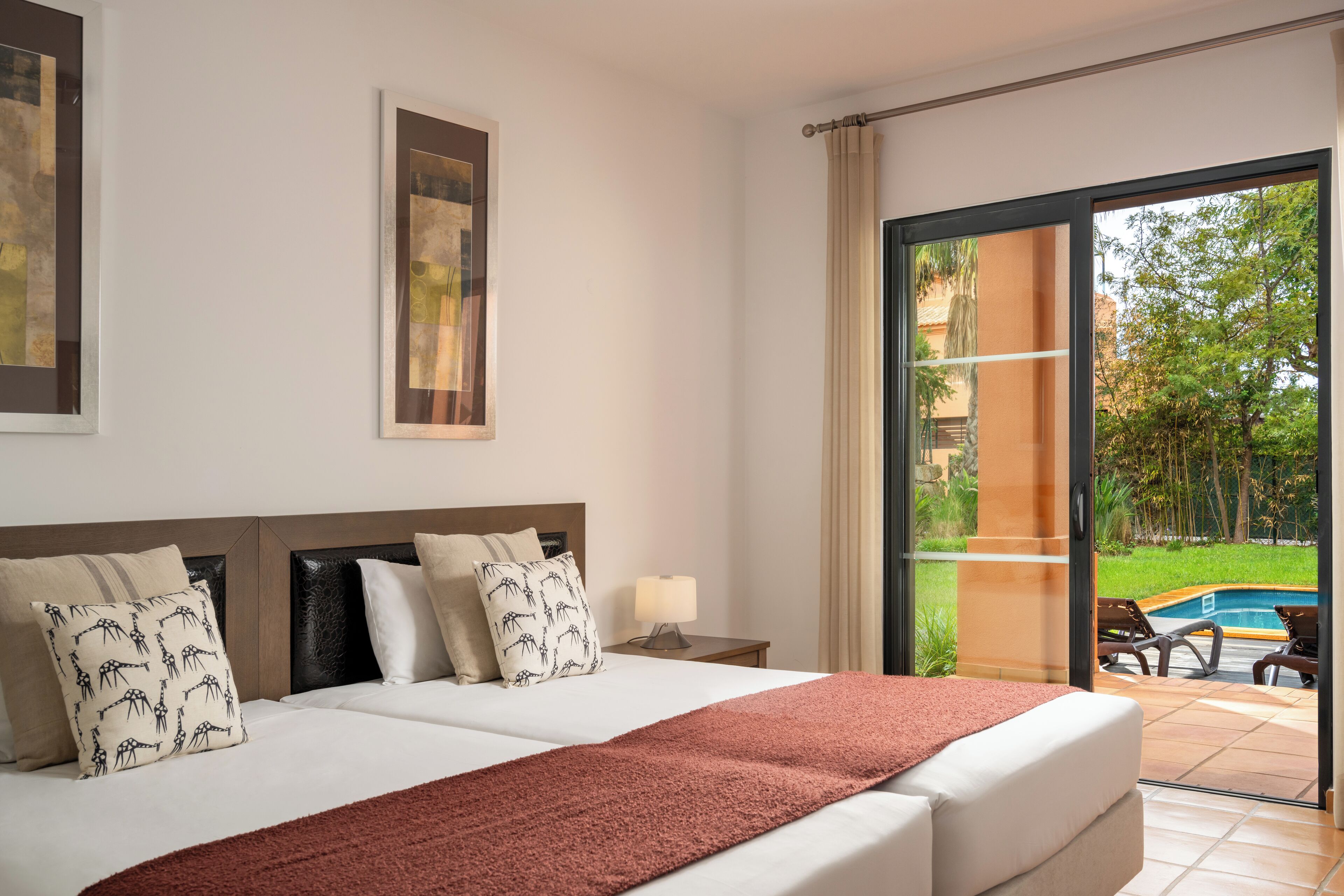 Een hotelkamer met een groot bed, twee kussens, een nachtkastje met een lamp en uitzicht op een zwembad en groen buiten.