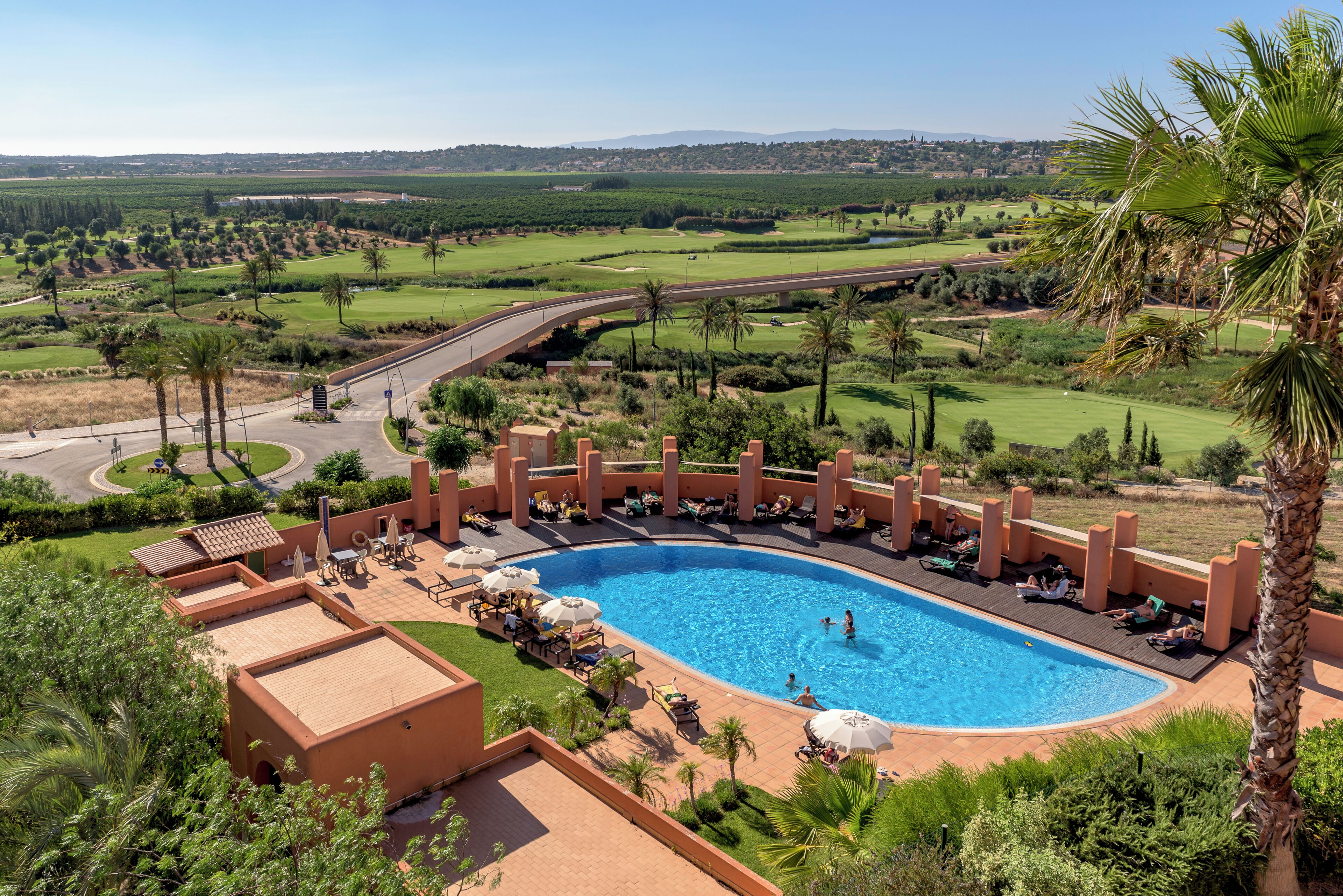 Un resort con piscina rodeada de sillones de descanso y sombrillas, con campos de golf y palmeras al fondo.