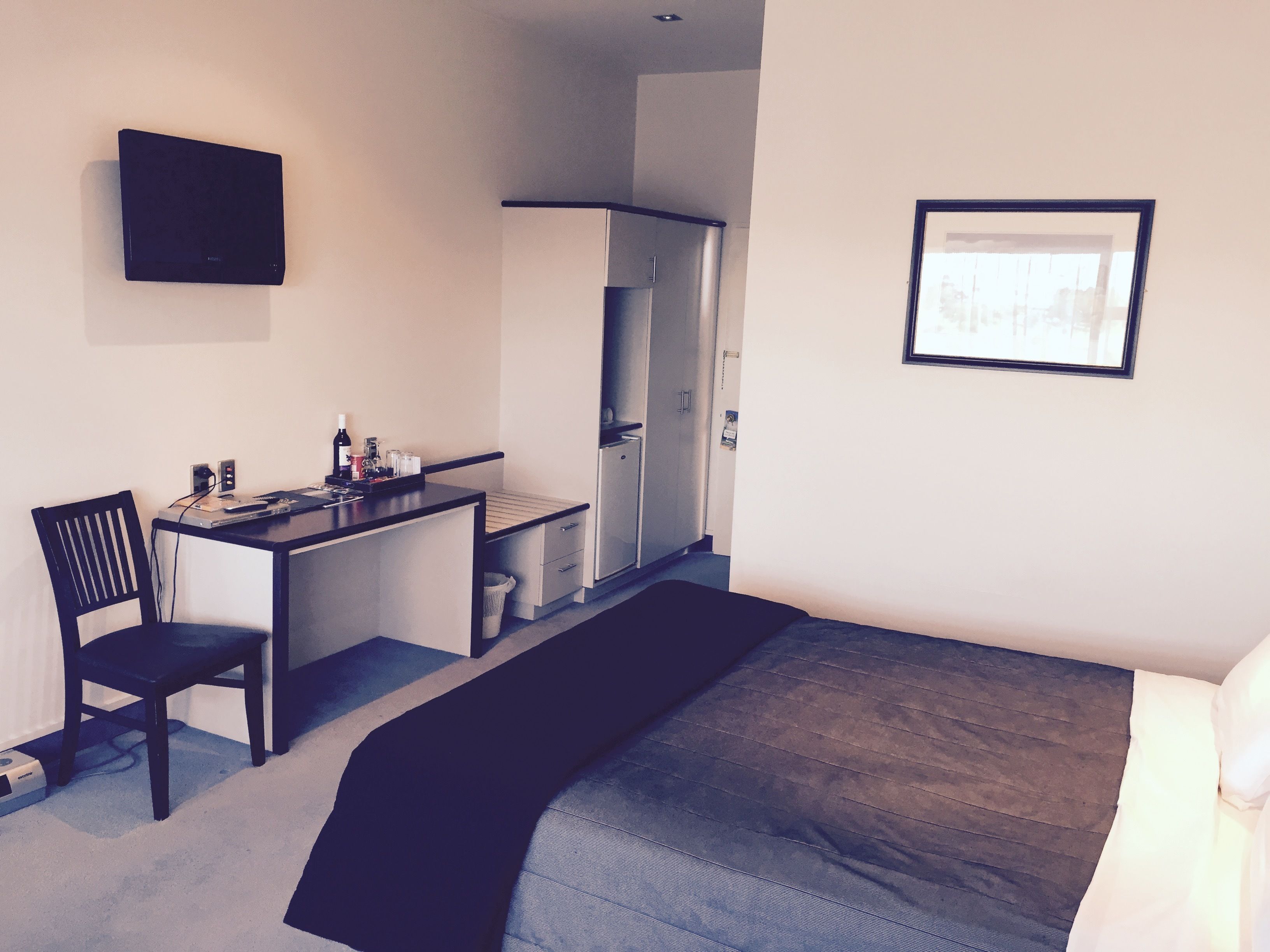 Quarto de hotel pequeno com uma cama, uma escrivaninha, uma cadeira, uma televisão e um quadro na parede.