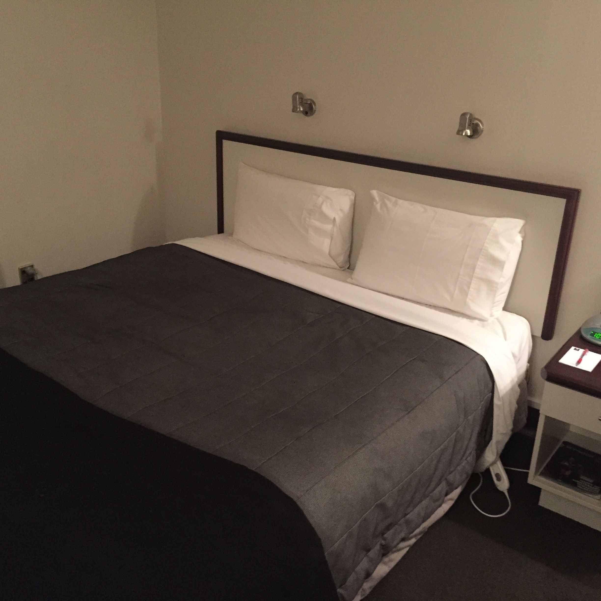 Una cama bien hecha con una colcha gris oscuro y almohadas blancas, una mesita de noche con un reloj y un enchufe en la pared.