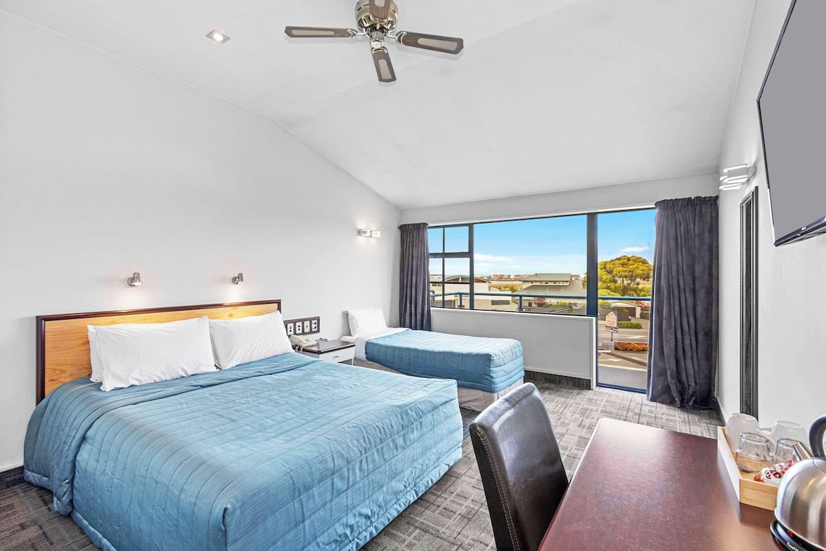 A hotel room with two beds, a dining table, a ceiling fan, and a large window with a view.