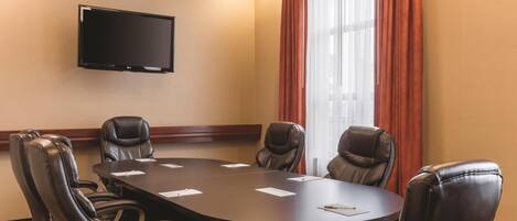 A conference room with a large table, six leather chairs, a flat-screen TV on the wall, and a window with red curtains.