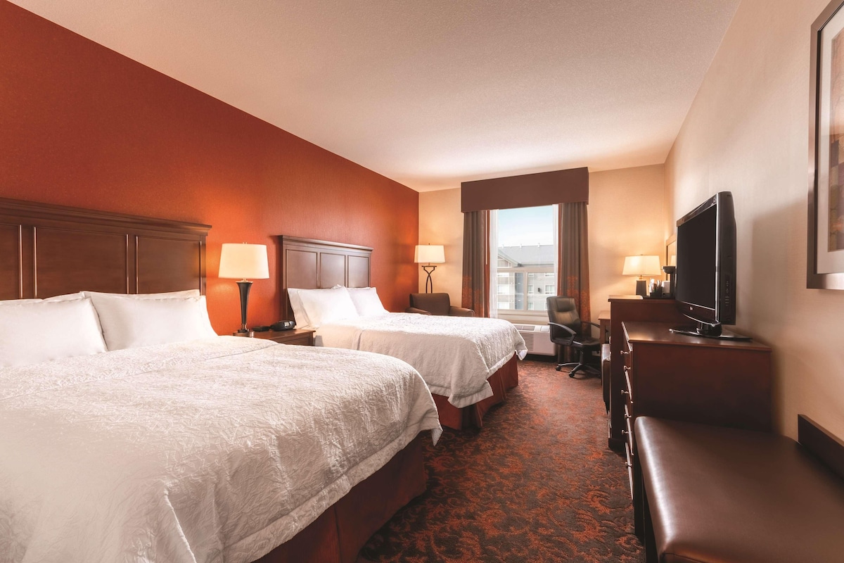 A hotel room with two beds, a desk with a television, and a view of the city through a large window.