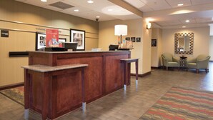 A hotel reception area with a wooden reception desk, a check-in counter, a framed welcome sign, and a patterned carpet.