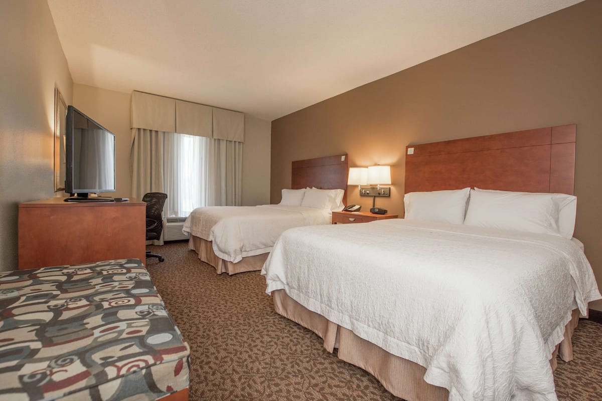 A hotel room with two beds, a desk, a TV, and a window with curtains.