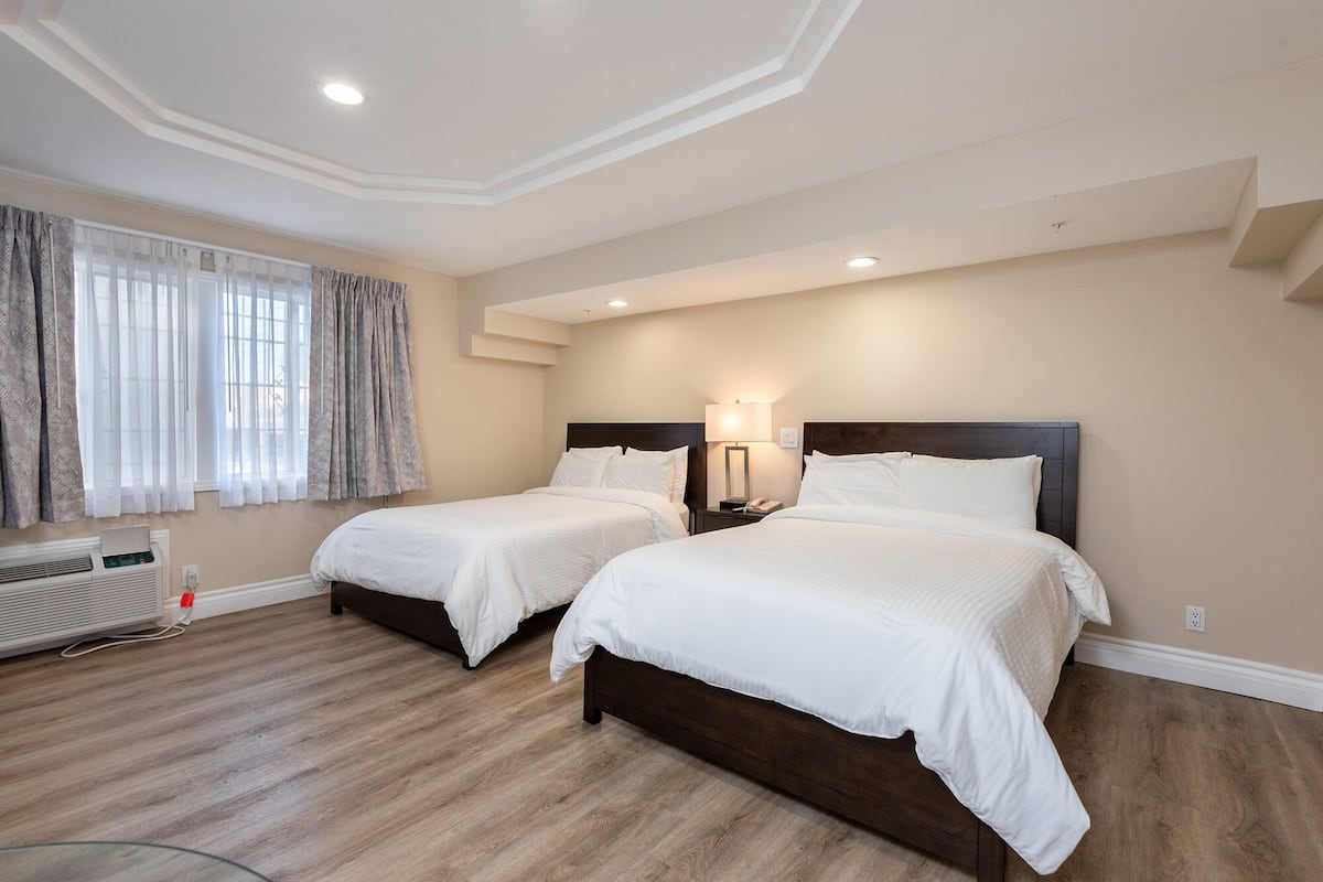 Habitación de hotel con dos camas, una mesita de noche con lámpara, una ventana con cortinas y un radiador.