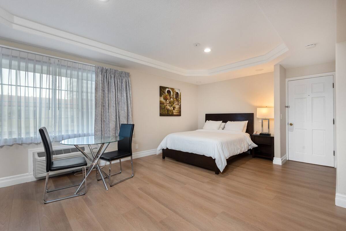Habitación de hotel moderna con cama, mesita de noche, lámpara y una silla cerca de una ventana con cortinas translúcidas.