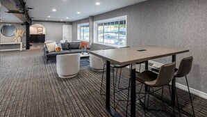 Una habitación de hotel moderna con un ventanal grande, un comedor con mesa y sillas, un sofá y un pequeño rincón de estar con un banco y una mesa redonda.