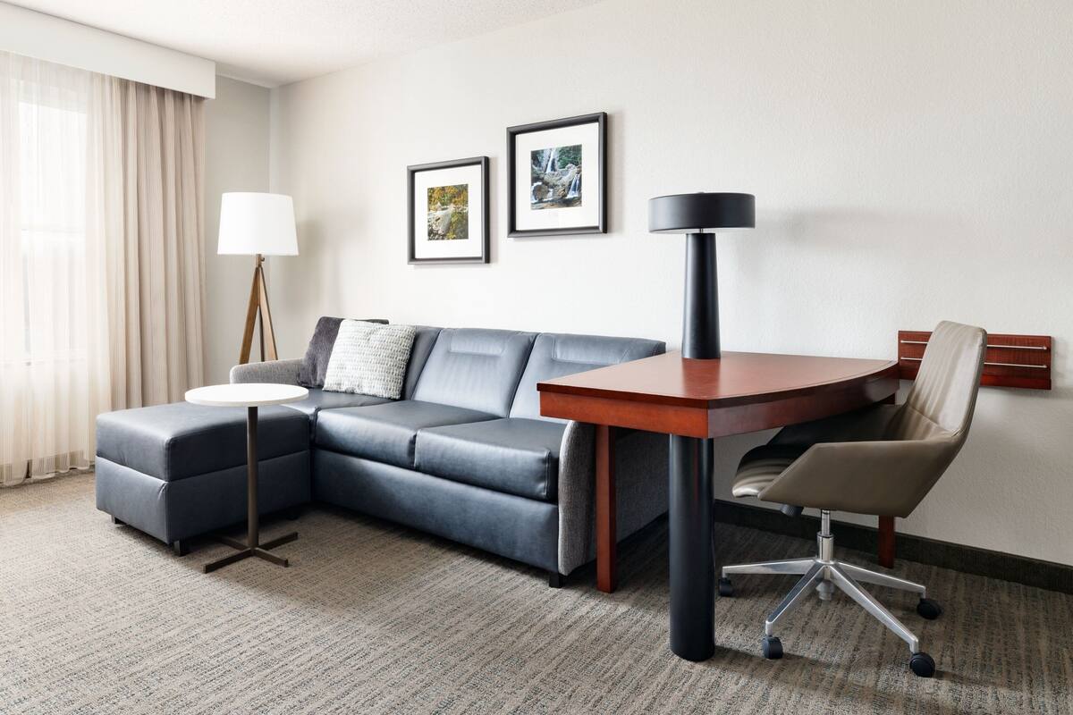 A hotel room with a grey sofa, a small round table, a desk with a chair, and two framed pictures on the wall.
