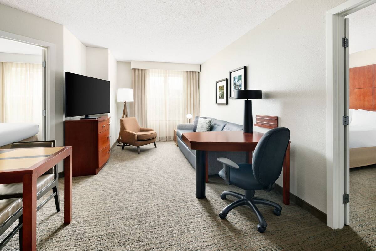 A hotel room with a bed, a desk with a chair, a TV, a sofa, and a small table.