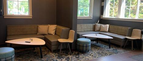 A modern living room with a sectional sofa, a round coffee table, and a small ottoman. There are two windows providing natural light and a view of greenery outside.