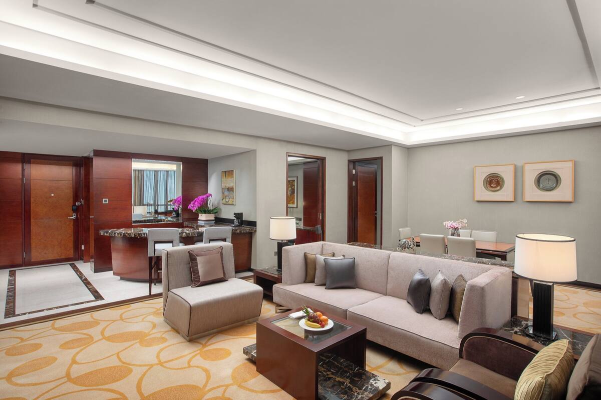 Ein modernes Hotelzimmer mit Sofa, Sessel und Couchtisch mit Obst. Es gibt eine Küchenzeile mit Arbeitsfläche und Spüle. Der Raum hat Holztüren und ein großes Fenster mit Vorhängen.