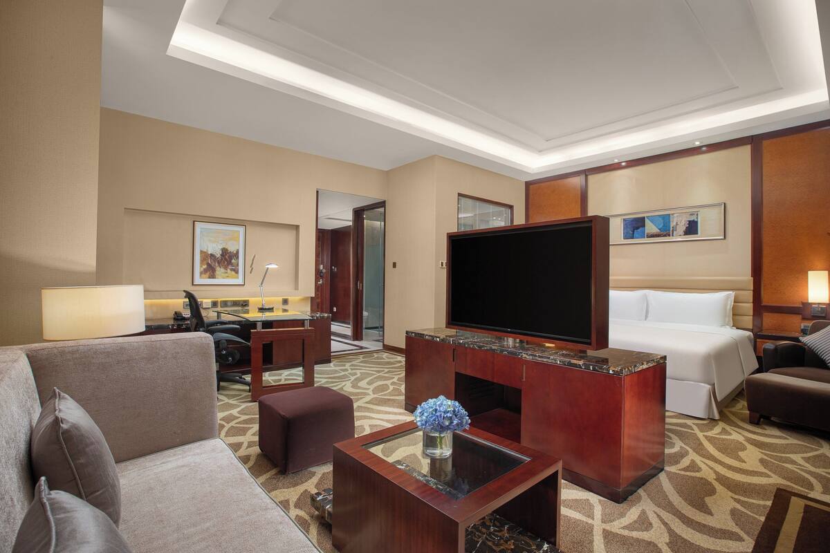 Ein Hotelzimmer mit großem Flachbildfernseher, einer Couch, einem Couchtisch mit Vase, einem Bett und einem Schreibtisch mit Lampe.