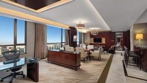 Habitación de hotel moderna con un escritorio de vidrio, una zona de estar, un bar y un ventanal con vistas a la ciudad.