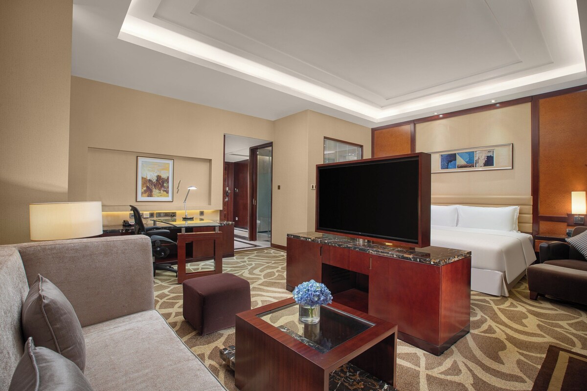 Habitación de hotel con una amplia televisión de pantalla plana, un sofá, una mesa de centro con un jarrón, una cama y un escritorio con una lámpara.