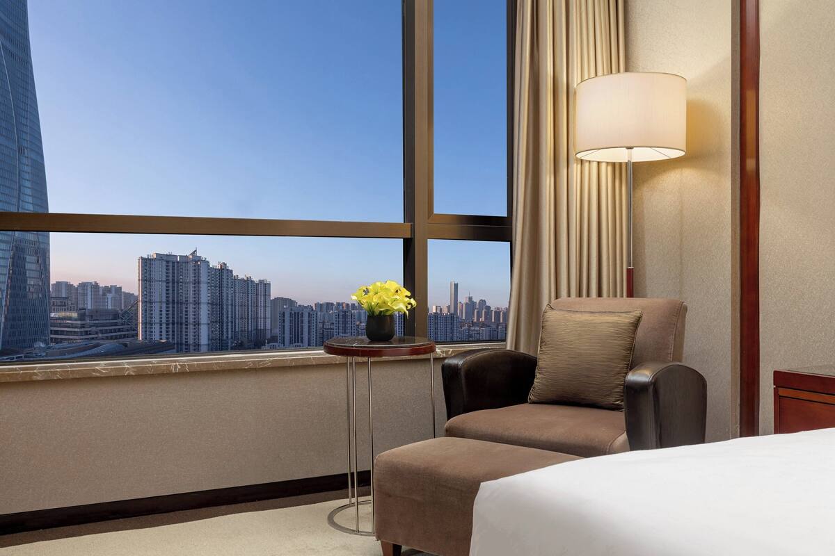 Ein Hotelzimmer mit großem Fenster, einem Bett, einem Sessel, einem Tisch mit Blumenvase und einer Lampe.