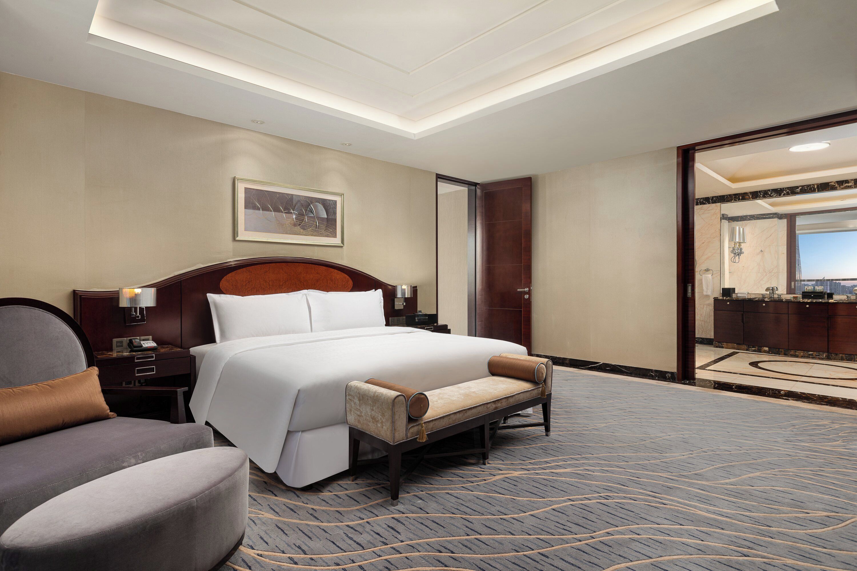 Una habitación de hotel con una cama grande, un banco, una silla, una mesita de noche, una lámpara y un cuadro en la pared.