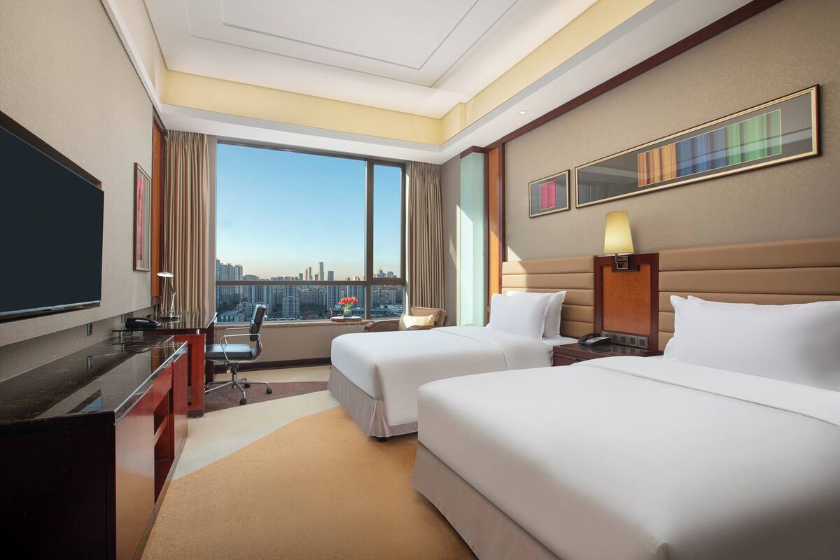Ein Hotelzimmer mit zwei Betten, einem Schreibtisch, einem Fernseher und einem großen Fenster mit Blick auf die Stadt.