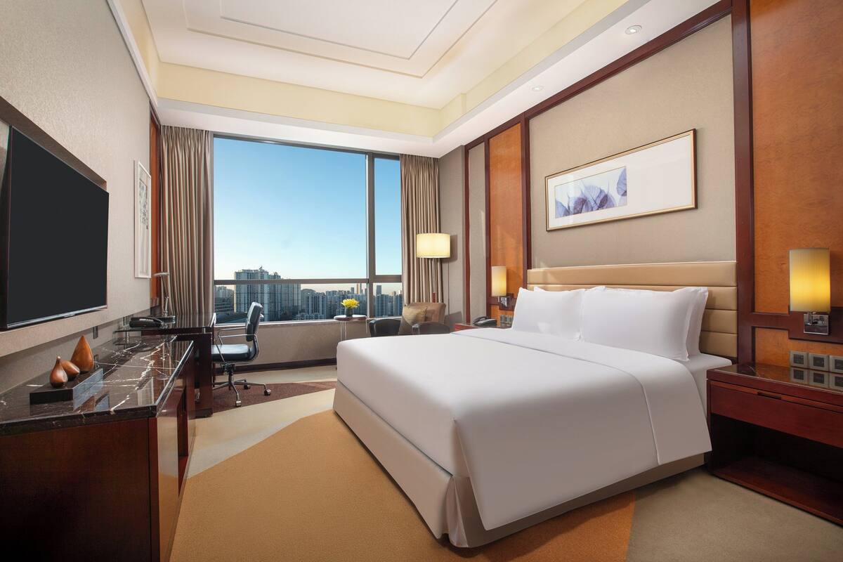 Ein Hotelzimmer mit einem großen Bett, einem Schreibtisch, einem Stuhl, einem Fernseher und Blick auf die Stadt.