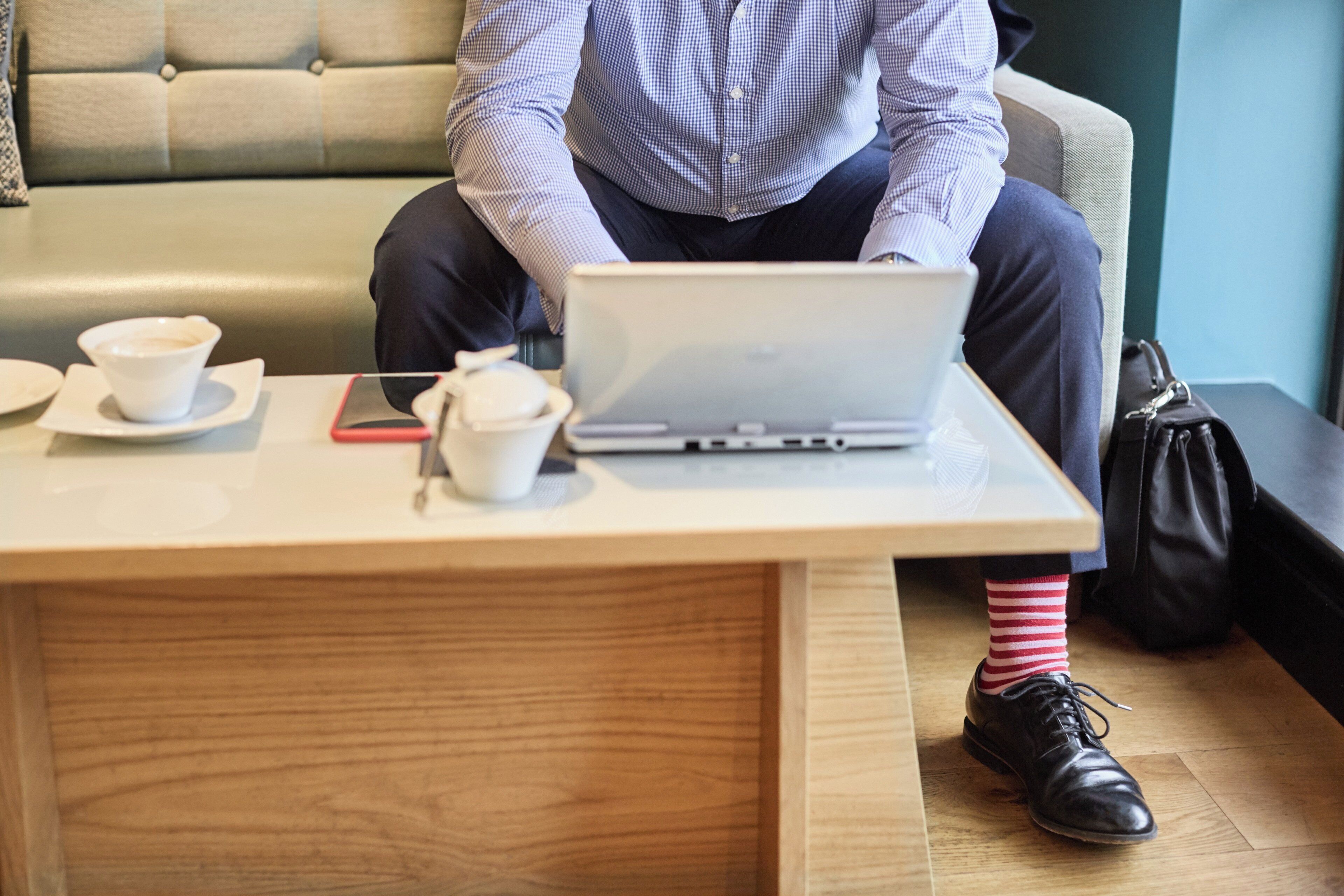 A person seated at a table with a laptop, a smartphone, and two cups, likely in a cafe or waiting area.
