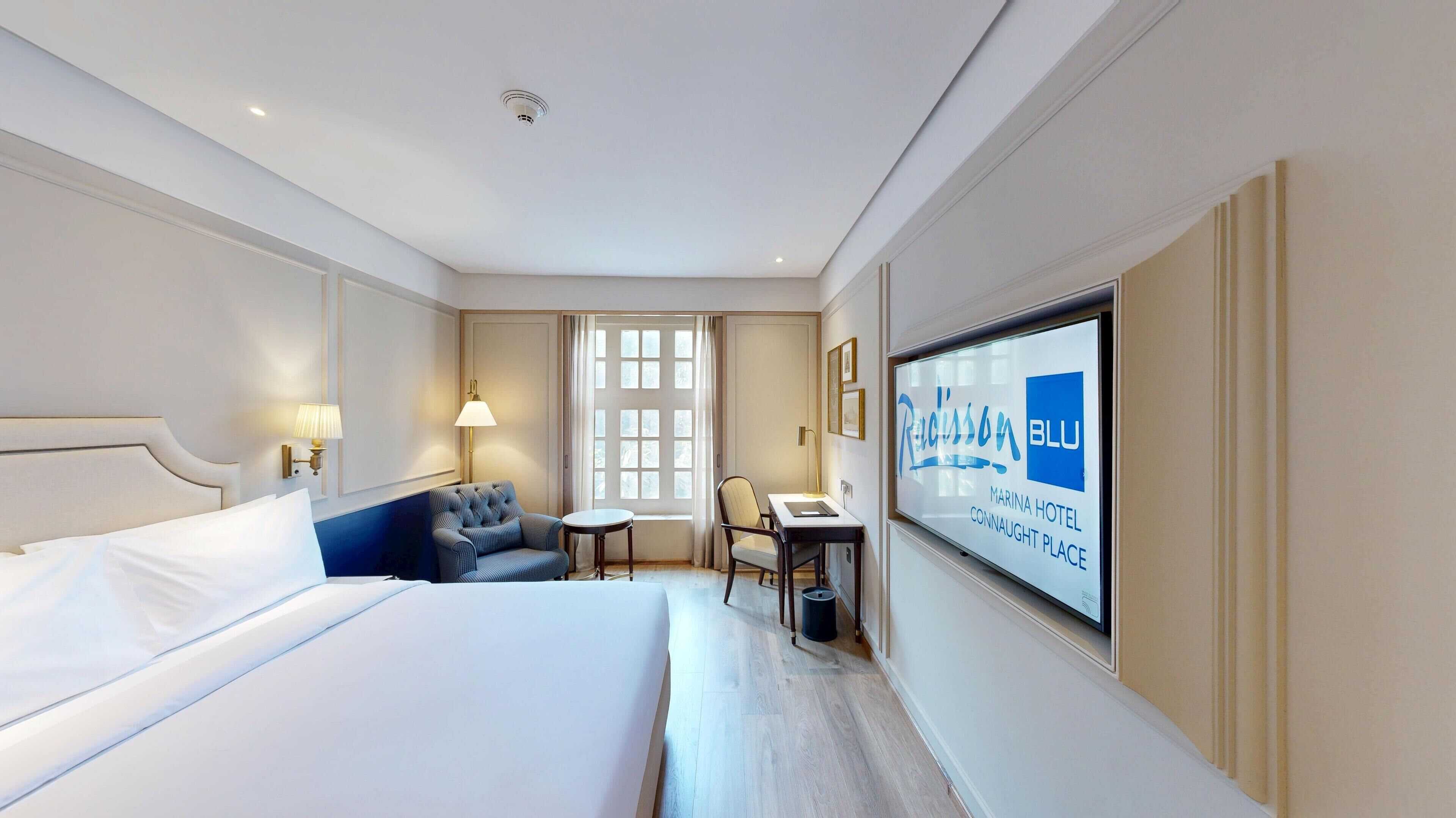 A hotel room with a bed, a desk, a chair, and a TV displaying the hotel's name.