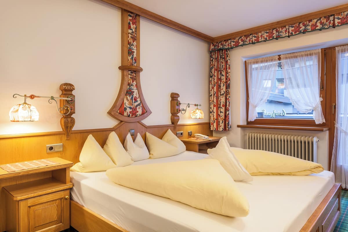 Una cama bien hecha con sábanas y almohadas blancas, una mesita de noche de madera con una lámpara y una ventana con cortinas estampadas.