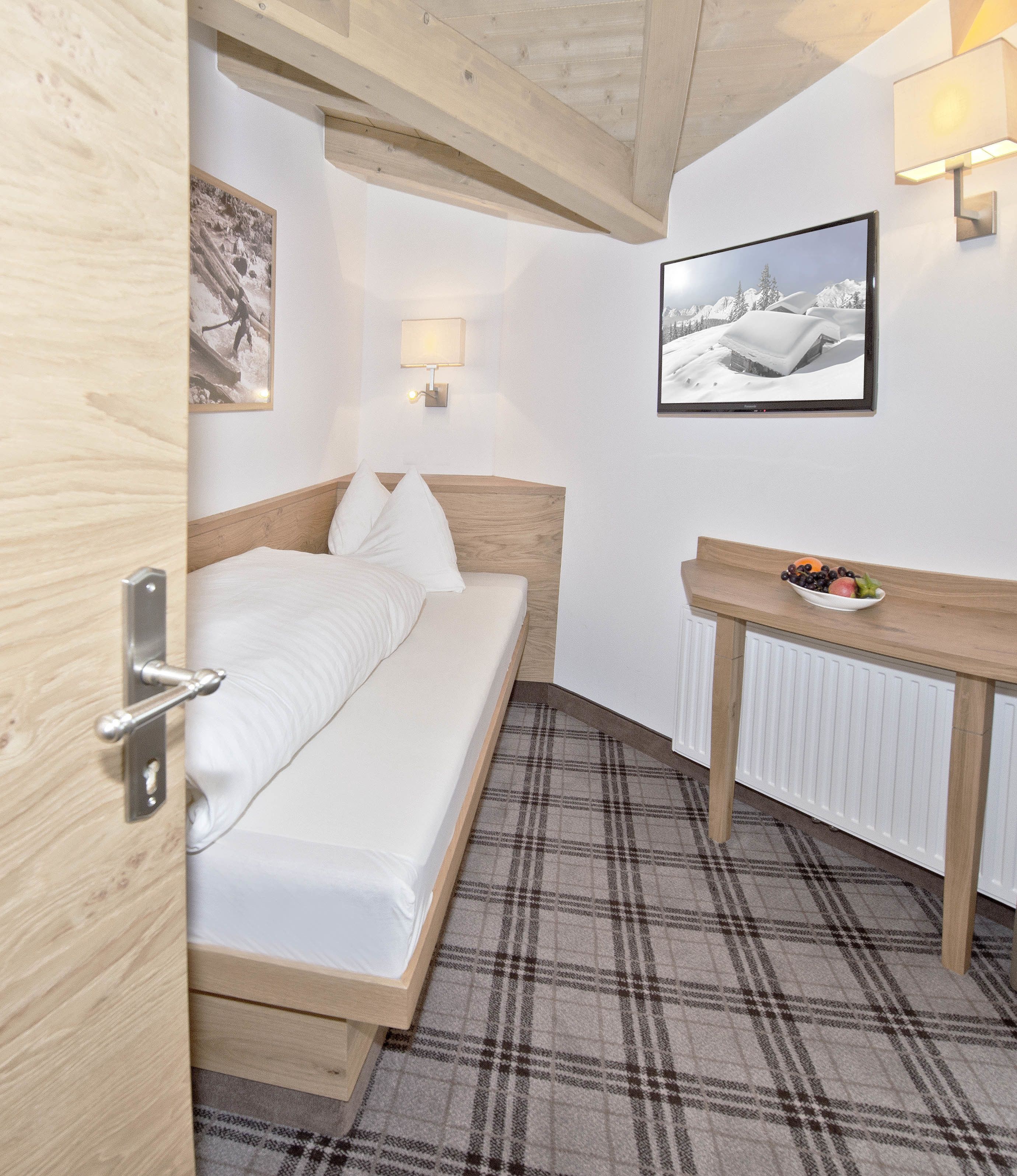A bedroom with a bed, a bedside table with a bowl of fruit, a wall-mounted lamp, and a framed picture.