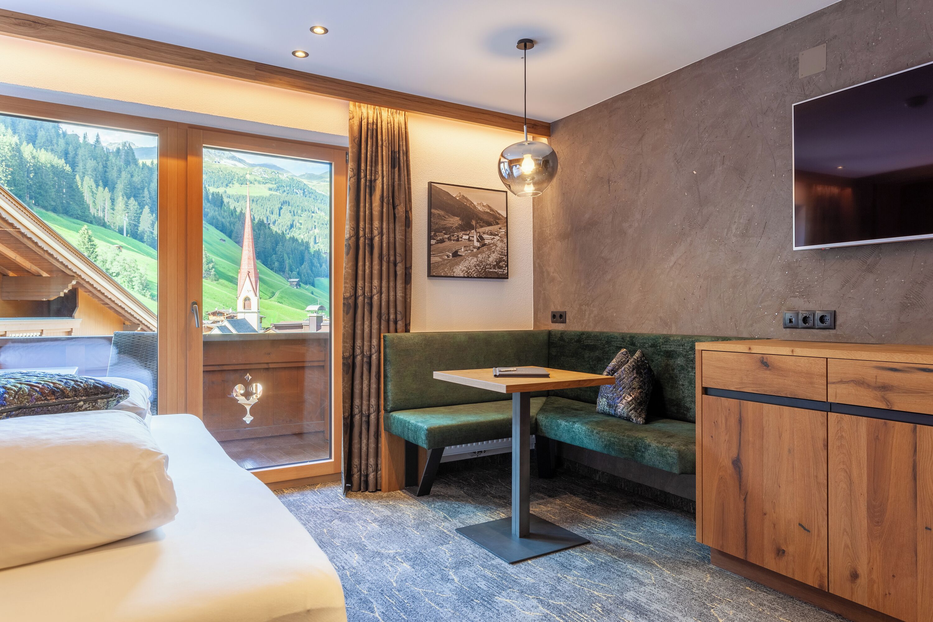 A room with a bed, a green sofa, a small table, a TV, and a large window with a view of a church and mountains.