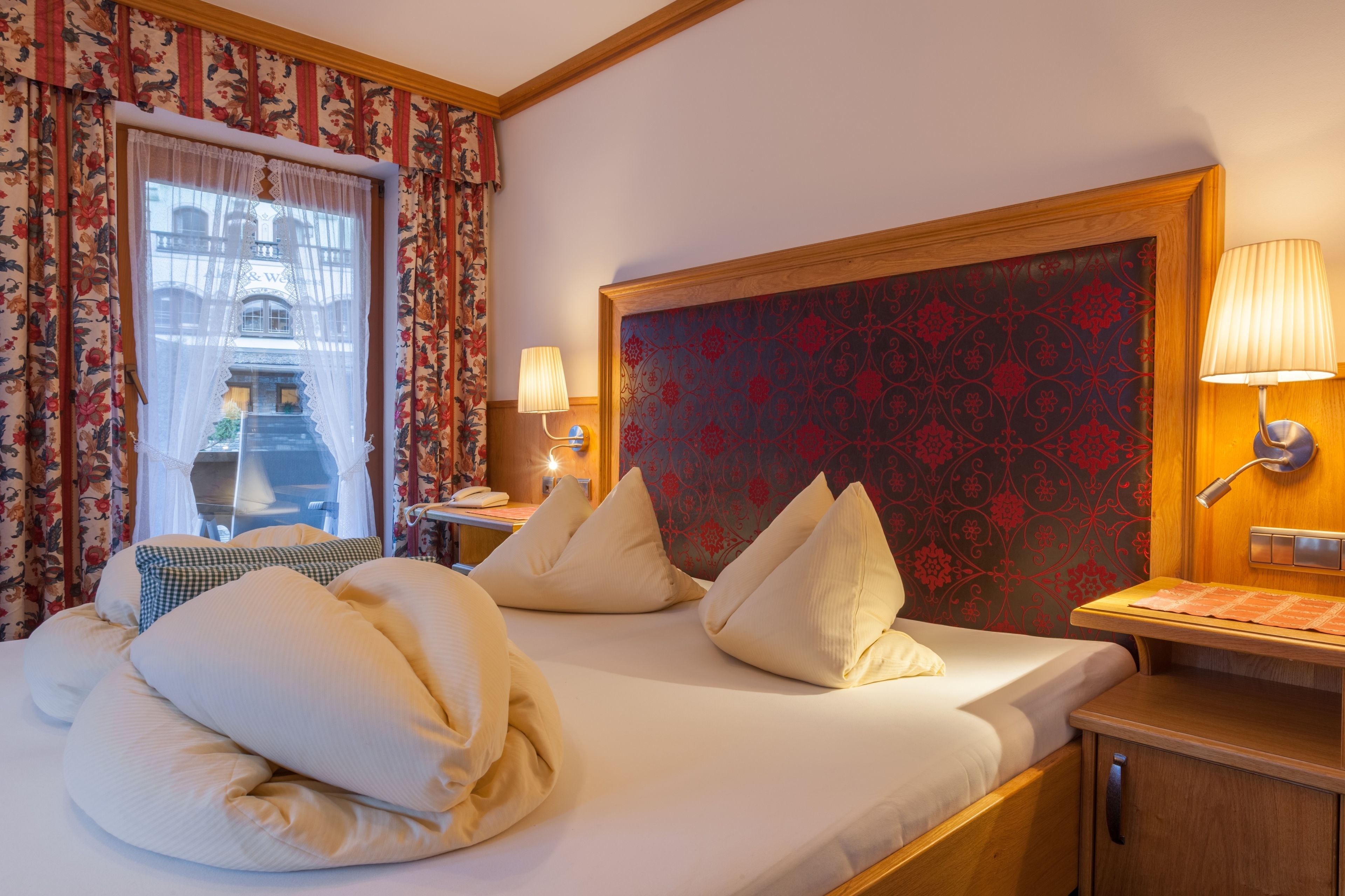 A hotel room with a large bed, floral curtains, a patterned headboard, bedside lamps, and a view of a building through the window.