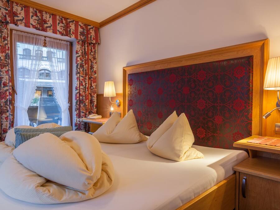 Quarto de hotel com uma cama grande, cortinas florais, cabeceira estampada, abajures e vista para um edifício pela janela.