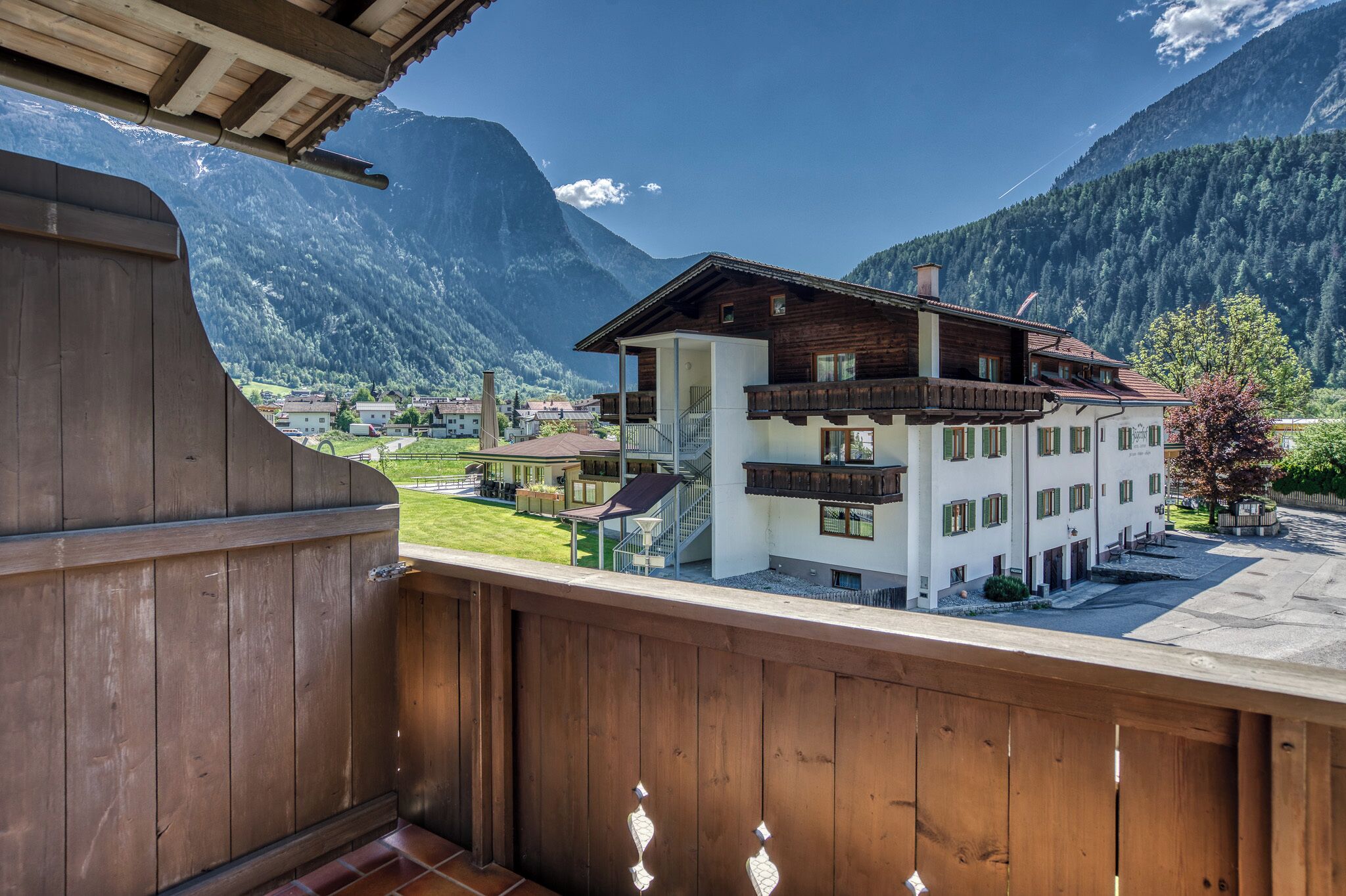 A wooden balcony overlooking a mountain village with a white building featuring balconies and a staircase.