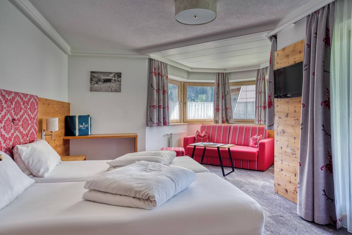 A hotel room with two beds, a red sofa, a wooden headboard, a TV, and a window with curtains.